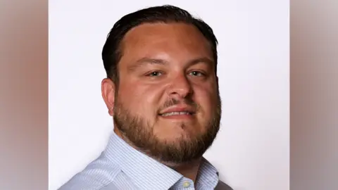 Essex County Council Sam Journet wears a blue striped shirt, facing to the right of the frame, but his head is turned to the camera. He has dark hair and a dark beard.