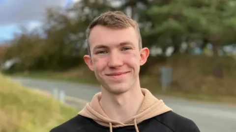 Isaac, a young man wearing a beige hoodie with a black jumper, with short dark blonde hair, smiles at the camera