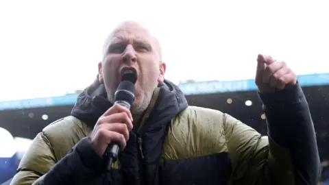 A bald man wearing a green and black raincoat talks into a microphone.