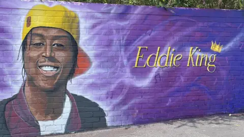 A close up of a mural showing Eddie Kinuthia smiling, wearing a yellow cap and black jacket. Beside him is scrawled writing, which reads 'Eddie King' on the right and 'talent, godsent, I'm chosen' on the left, with various music notes against a bright purple background.