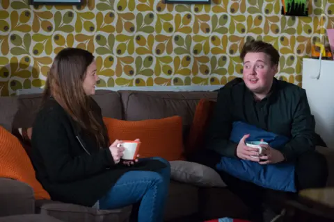 Lacey Turner who plays Stacey Slater (left) and Riley Carter Millington, who plays Kyle Slater (right). A woman with brown hair wearing a black jumper and blue jeans sits on a sofa holding a mug next to a man in a black shirt holding a mug.