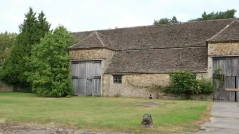 Large tiled roof barn with two sets of double barn doors. Between these doors, the roof profile is lower than the height of the doors. A small grass lawn can ben seen in front of the barn, with a concrete path leading to one of the doors.