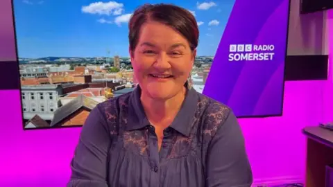 Emma Britton. She has short dark hair and is wearing a blue shirt. She is pictured in the BBC Radio Somerset studio, in front of a purple wall and a screen showing a landscape photo of the county.