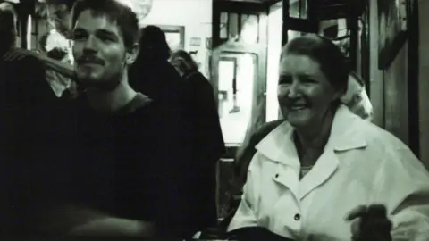 Glen Grundle A black and white effect photograph of a younger Glen with his mother. They are sat next to each other smiling and looking to the side of the camera
