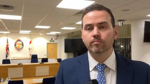 A man in his 30s with short hair and a trimmed beard looking towards the camera. He's wearing a blue suit and tie with a white shirt. Behind him are desks and chairs in a council building. At the back out of focus is a coat of arms with the union jack flag on the left and St George's flag on the right.
