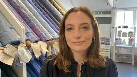 Natalie Mumford looking at the camera. She has shoulder length brown hair and is wearing a black blouse. Behind her is a row of purple patterned silk on a white shelf. 