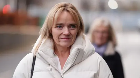 PA Media Sandie Peggie - a woman in a white jacket with blonde hair - walking down a street. She is smiling.