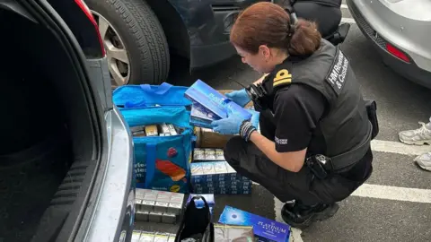A female HMRC officer kneeling down and examining boxes of cigarettes and tobacco. She is dressed in black, with blue plastic gloves on.