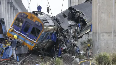 Rescuers work the wreckage of a passenger train after a construction crane collapsed onto it. The train is partially suspended, with parts of it completely crushed