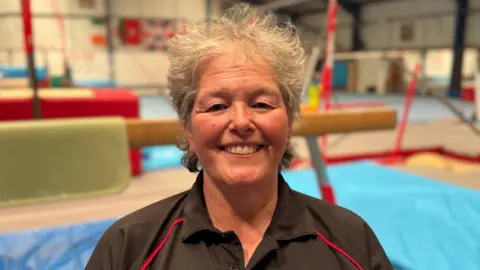 Helen Duggan smiles she wears a black t-shirt and has short grey hair. She stands in a gymnasium in front of equipment such as brown balance bar.