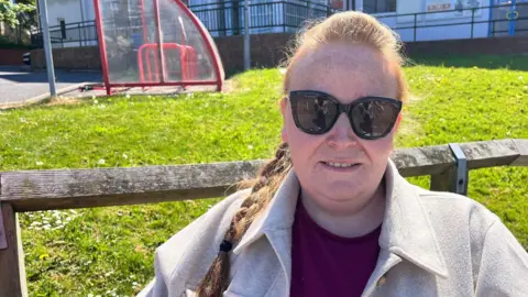 A woman with ginger hair, tied in a pleat, sits in the grounds of a leisure centre in bright sunshine. She is wearing black sunglasses, a cream-coloured coat and a claret-coloured top. Behind her is a wooden fence, a green lawn and a red-framed glass bike shelter.