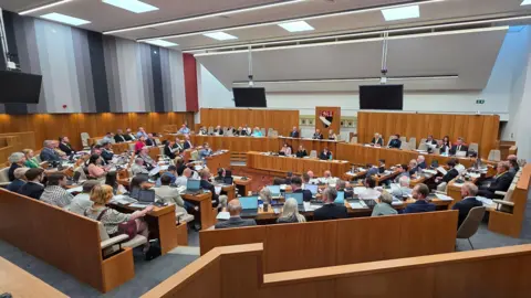 Paul Moseley/BBC The airy wood-panelled council chamber at Norfolk County Council with around 60 councillors sitting in a crescent looking up at a raised platform on which another 10 senior councillors and officials are sitting. 
