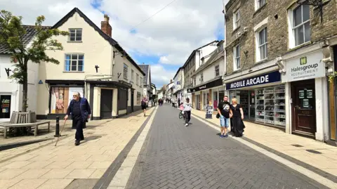 The main street in Diss, Mere Street. It's a sunny day with shops on either side of a pedestrianised area. There are a few shoppers walking in the street.