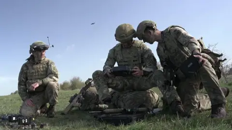 Three soldiers in camoflage gathered around a hand-held computer while a drone flies overhead