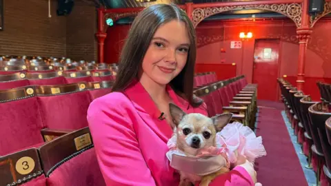Macie sits in a row of theatre seats holding a chihuahua dog that is wearing a pink tutu. Macie has long brown hair and is wearing a pink jacket 