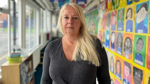 BBC Helen Slack has blonde hair and is wearing a grey long sleeve top. She is standing inside the school with drawings on walls to her right