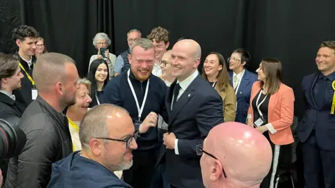 Stephen Flynn seen in a group of people celebrating his win, being congratulated by a man with a beard.