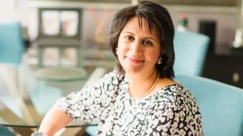Handout photo of Mrs Virani sat on a blue chair around a glass table. She is smiling at the camera, with short brown hair and earrings in. She has a black top on with different coloured patterns across it. 