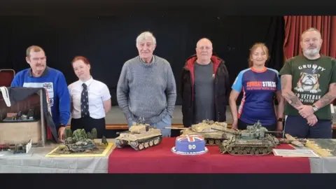 Veterans and Community hub A group of people standing in front of a table with a numerous items on it. There are models of tanks and a blue, red and white cake with the number 80 on it.