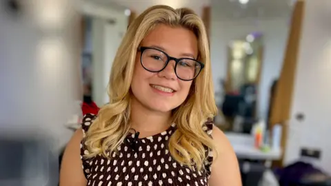 Jessica Flaxman stands in a hair salon with the background blurred out. She has long blonde hair and wears black-rimmed glasses. She is dressed in a brown dress with white polka dots.