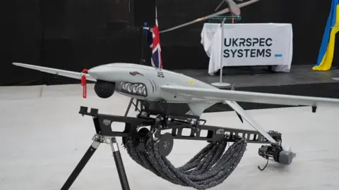 A Ukrspecsystems drone with a shark face and design on its nose propped up on a black metal stand. In the background there is a white banner which reads Ukrspecsystems and a Ukraine flag.