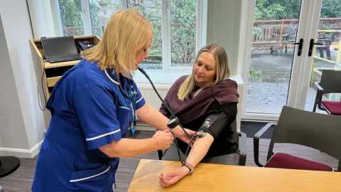 A 45-year-old woman is sitting at a table in a room and having her blood pressure taken by a nurse. The nurse is wearing a blue uniform and is listening to the woman's pulse through a stethoscope.
