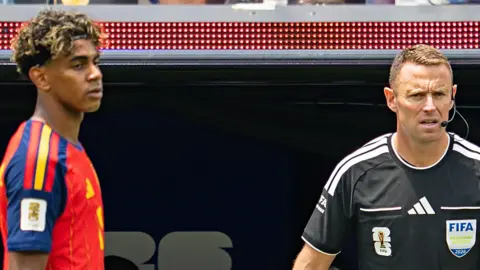 A close up photo of Stuart Burt in a black referee top with an earpiece and microphone. On the other side of the picture is Spanish forward Lamine Yamal in a red Spanish football shirt. Both are looking at the action off camera.