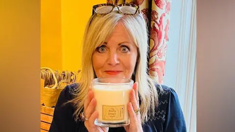 Supplied A photo of a woman holding a candle in front of her face. She is standing in front of a window with patterned curtains on it. Her eyes are wide and she is looking into the camera.