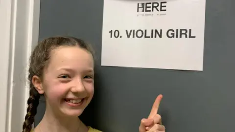 A teenage girl, 13, with French braids in her hair wearing a yellow top pointing at a sign on the back of the door that reads "10 Violin girl"