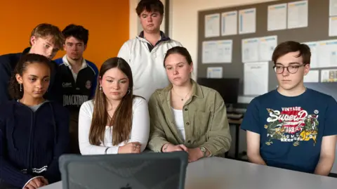Seven students, four boys and three girls are gathered around a computer in a classroom.