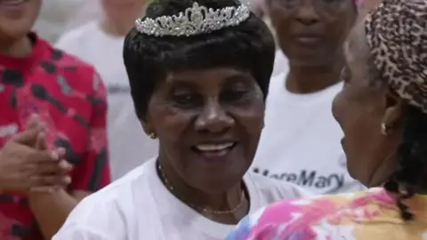 Mary Marson, a 95 year old black woman wearing a tiara, white t-shirt and smiling at the camera during a dance class