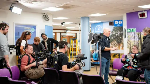 Cambridge University Hospitals A behind-the-scenes picture of the hospital waiting room at Addenbrooke's Hospital, with Shirley sitting in a purple chair knitting, while two camera crew are sitting in front of her, while another man is stood diagonally. Her daughter is standing on the side of her chatting to her.