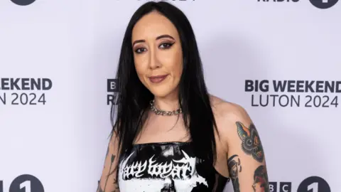 Getty Images Alyx Holcombe wearing a black top with white writing on it. She has tattoos on both arms, black hair and black jeans.