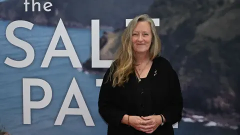 Salt Path author Raynor Winn pictured in front of a promotional book for her novel. She has blonde long hair which is swept onto her one shoulder and smiles at the camera.