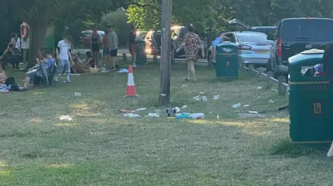 Peter Markham Rubbish is strewn across a village green, with empty cups and bottles visible on the grass. About 20 people mill around or sit on the grass. There are several cars parked on the adjacent road.