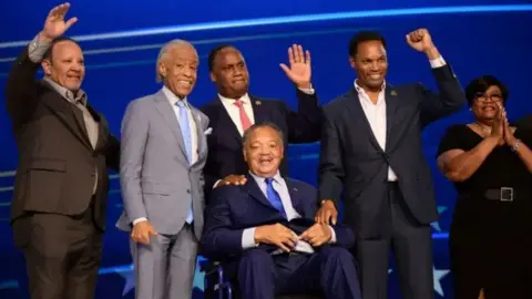 Getty Images Jackson is wearing a navy suit and tie and is sat in a wheelchair whilst five others are standing around him waving on stage during the first day of the Democratic National Convention in Chicago. 
