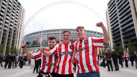 Three men in red and white striped Sheffield United shirts stand in front of Wembley stadium with their arms raised 