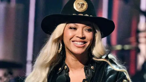 Getty Images Beyoncé on stage smiling as she receives an award. She's styled in a black cowboy hat with a gold medallion on the front and a black denim jacket. She has long platinum blonde hair and wears large gold hoop earrings. 