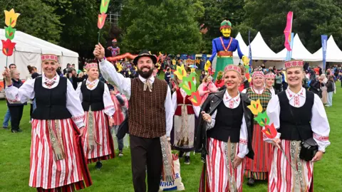 Pacemaker Group of people standing in traditional clothing with white festival tents behind them. 