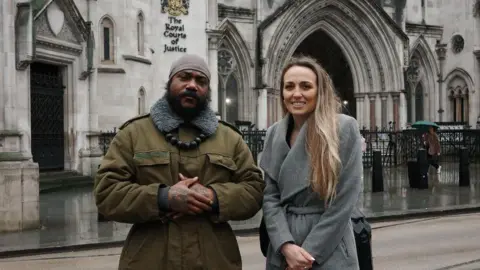 Big Brother Watch Shaun Thompson, a black man with a beard wearing a green coat and a large beaded necklace stands next to a white woman with long blond hair, wearing a grey coat. The pair are stood outside of The Royal Courts of Justice.