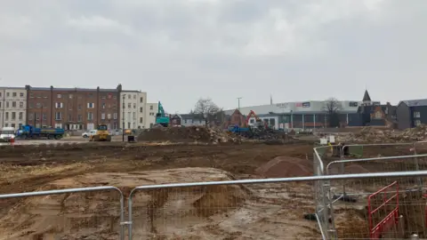 LDRS A brownfield site with mounds of dirt and rubble. Apartments and an industrial building can be seen in the background, with a road with several vehicles. There are metal fences in the foreground.