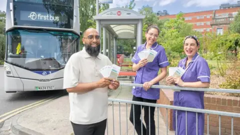 A First Bus representative and two female maternity staff stood on a pavement by a road. They are each holding travel tickets in their hands. Behind them is a parked double decker bus which reads 'First bus' on a digital screen.