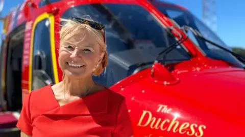 Cornwall Air Ambulance Barbara Sharples wearing a red dress and standing in front of a red Cornwall Air Ambulance. 