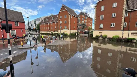 Zoie O'Brien/BBC A town centre junction where much of the road is underwater. There are people standing in the road on one of the dry sections.