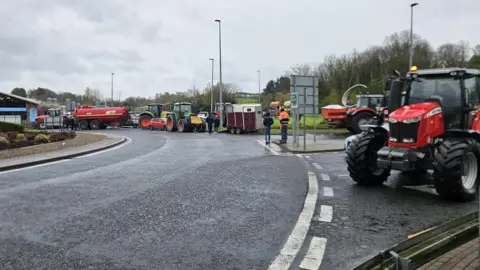 A road with trees in the background. The skies are cloudy. On the right are tractors, which appear to be at a standstill. There are tractors and a lorry-cab parked at the other side of the road. Some people are standing around.