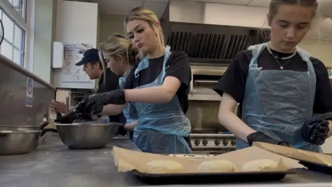 A man and three teenage girls in a kitchen standing over a work bench making scones.

The three girls with blonde hair are wearing black t-shirts and blue plastic aprons, 

There is a baking tray with dough in the shape of scones in the foreground