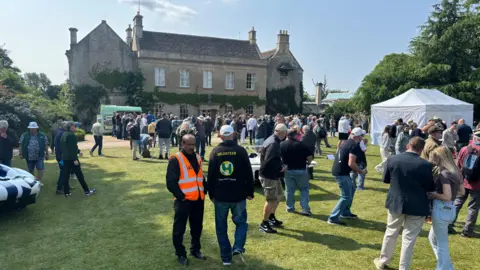 Middlewick House Open Garden A member of security wearing an orange hi-viz jacket and a volunteer can be seen in the foreground. There is a crowd of people behind them. The image is from last year's event.