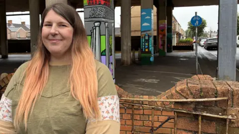 A woman with long blonde hair wearing a green top smiling in front of a wall and pillars that hold up the M4 with a road going under the motorway road deck