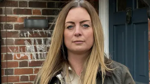 Claire Massey standing outside her home that was damaged by fire in Stoke-on-Trent. She has long light hair past her shoulders and wears a dark jacket over a light blouse. Behind her is a blue front door next to a bare brick wall with graffiti on it