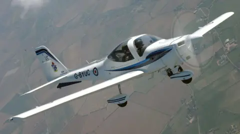 A small white RAF plane seating two people is seen mid-flight, with a view of fields below. 